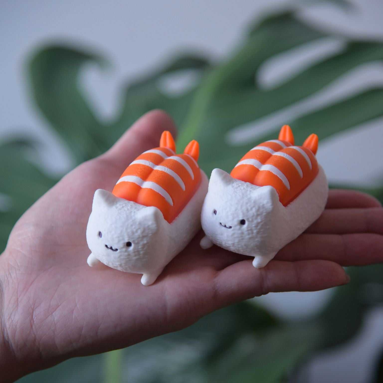 M.E.S.A. X Sushi Cat shrimp fidget clicker toy, Two small white cat figurines dressed as shrimp sushi with orange and white striped tails, resting on a person's open palm, featuring cute smiling faces and designed as keychain accessories.